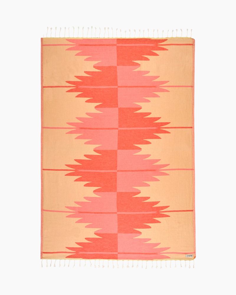 A vertically oriented image of a beach towel with a geometric pattern in shades of pink and orange, featuring decorative tassels at the ends.