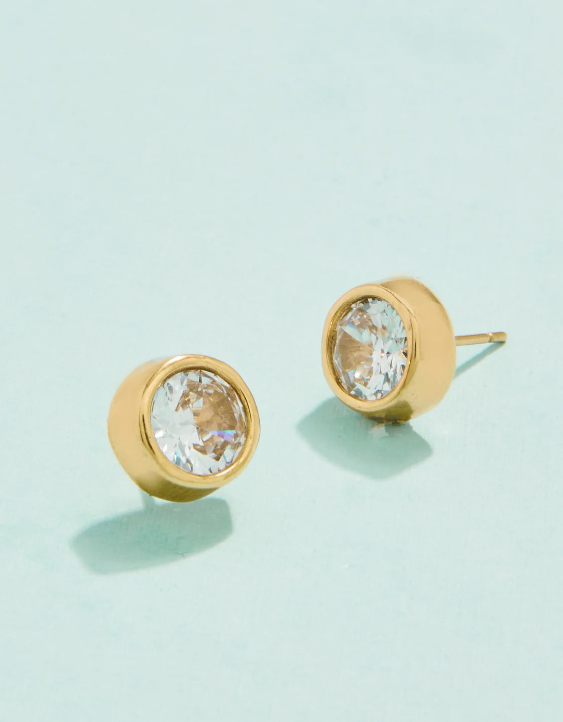 A pair of gold-colored stud earrings with cubic zirconia crystals on a light background.