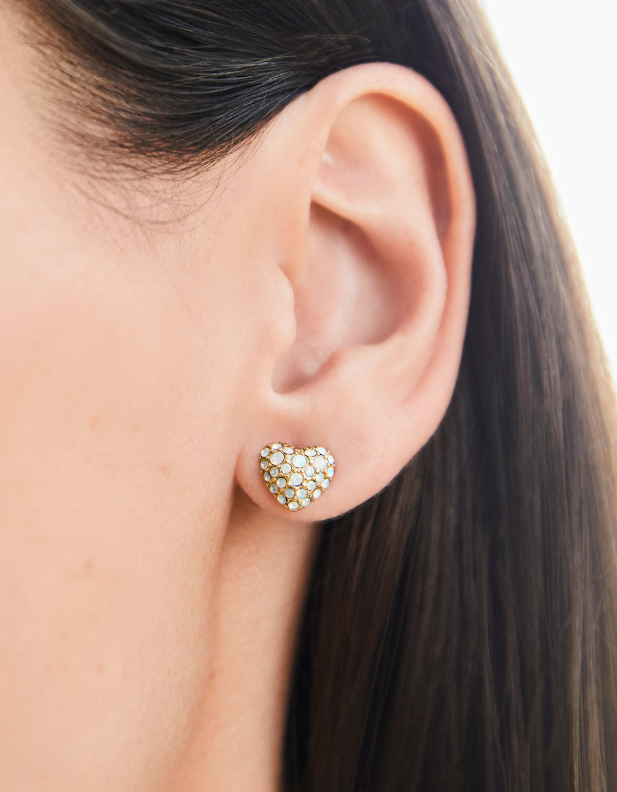 A close-up image of a woman's ear wearing a sparkling heart-shaped stud earring encrusted with white opal crystals.