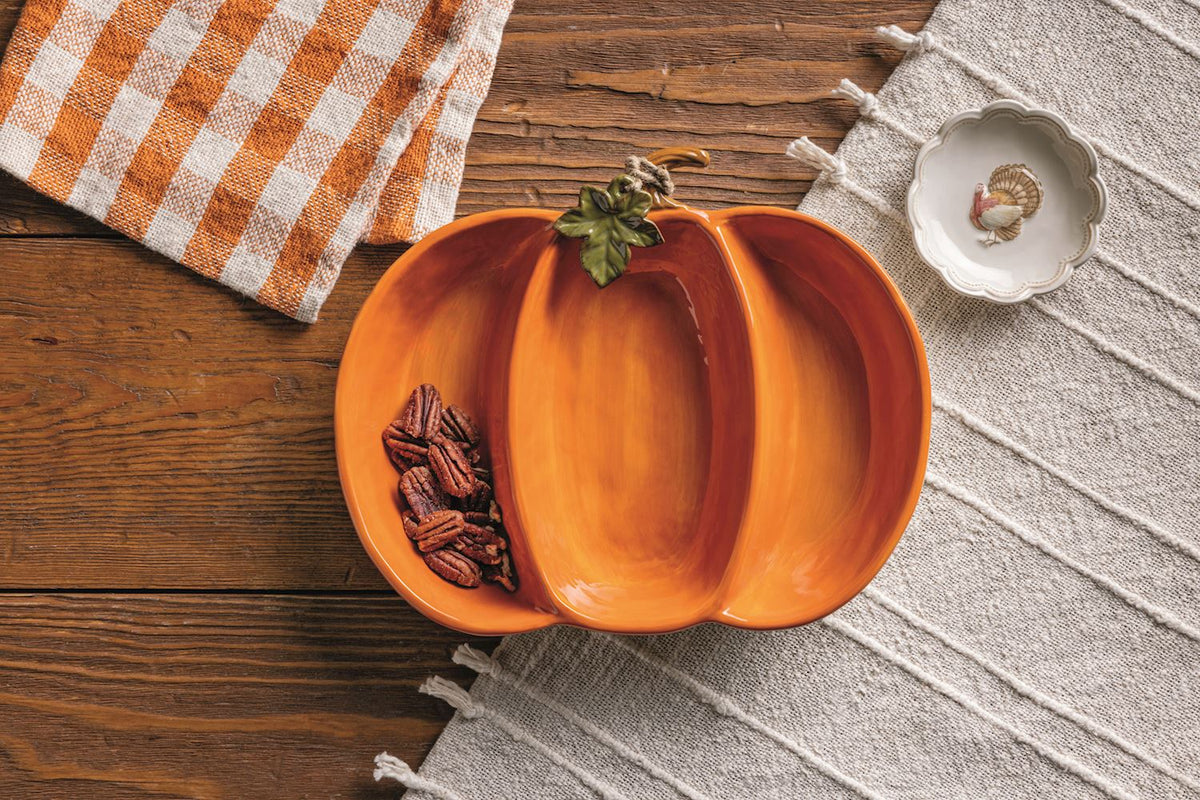 Pumpkin-shaped bowl with pecans on a wooden surface with a checkered cloth and gray textured mat.