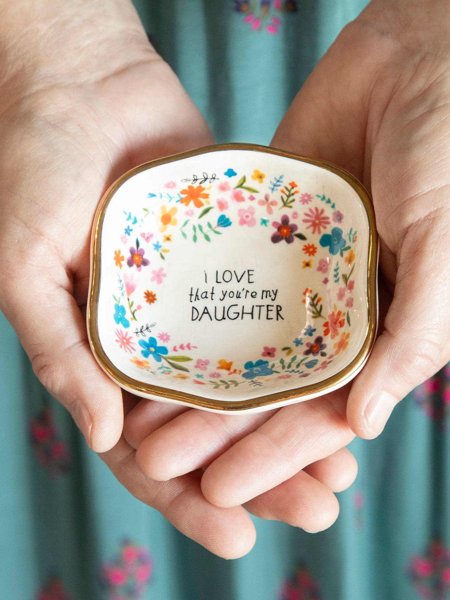 A hand holding an antiqued, floral-patterned trinket bowl with the inscription 'I love you, daughter' on it.