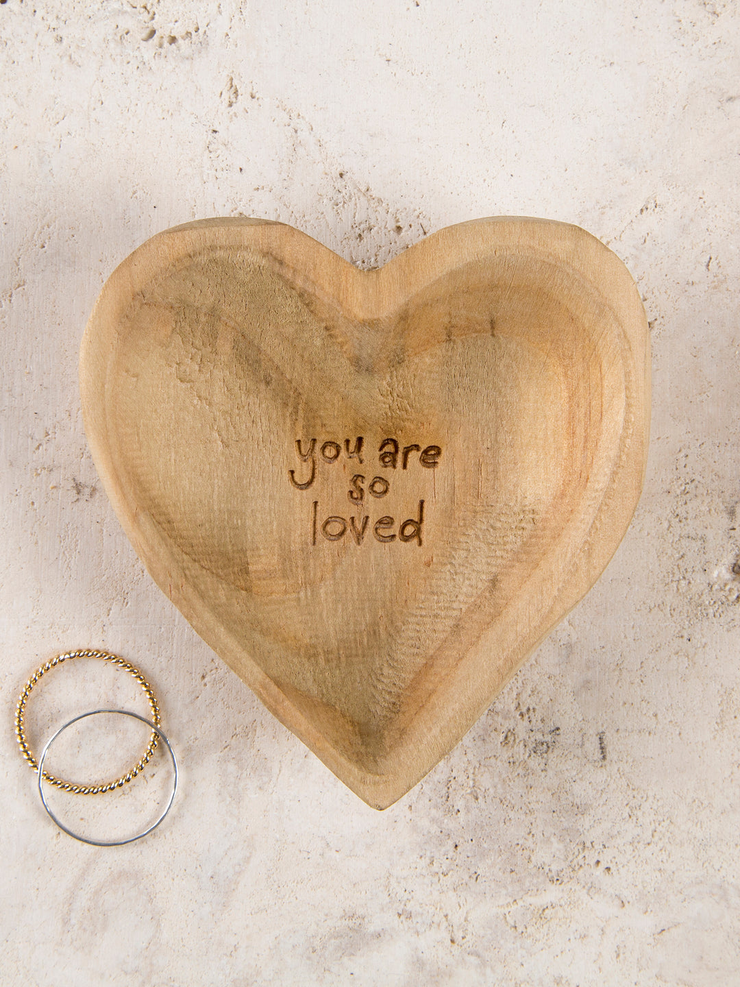 A wooden heart-shaped dish with the inscription 'you are so loved' on the inside, displayed with two rings beside it.