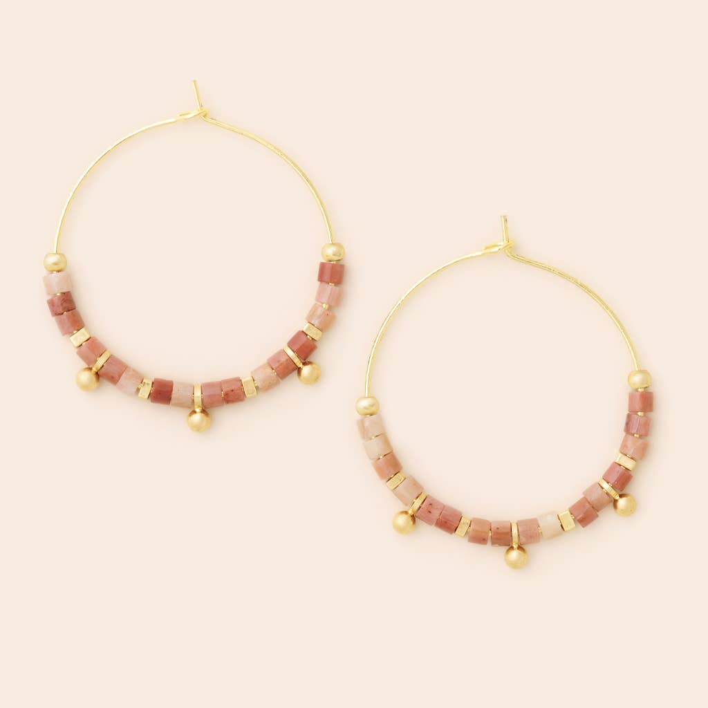 A pair of gold-colored hoop earrings with pink stone discs attached to the hoop, displayed against a light background.