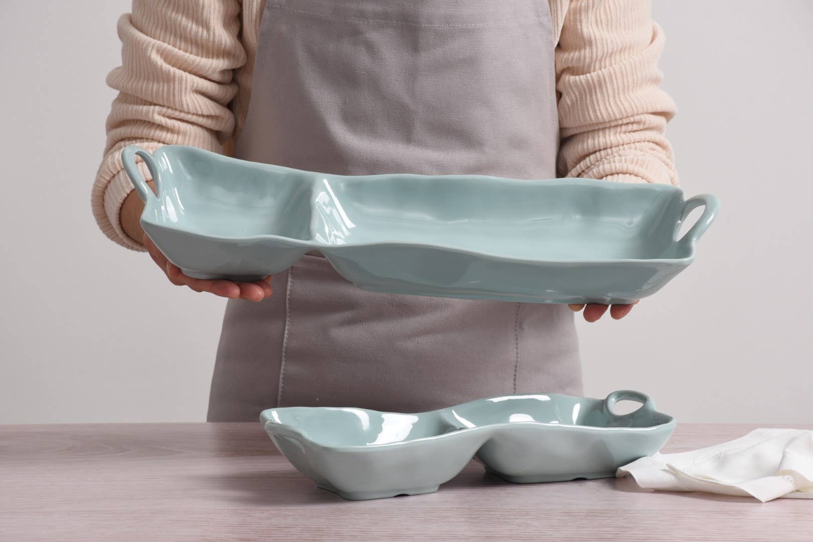 Person holding a light blue ceramic dish with handles on a neutral background
