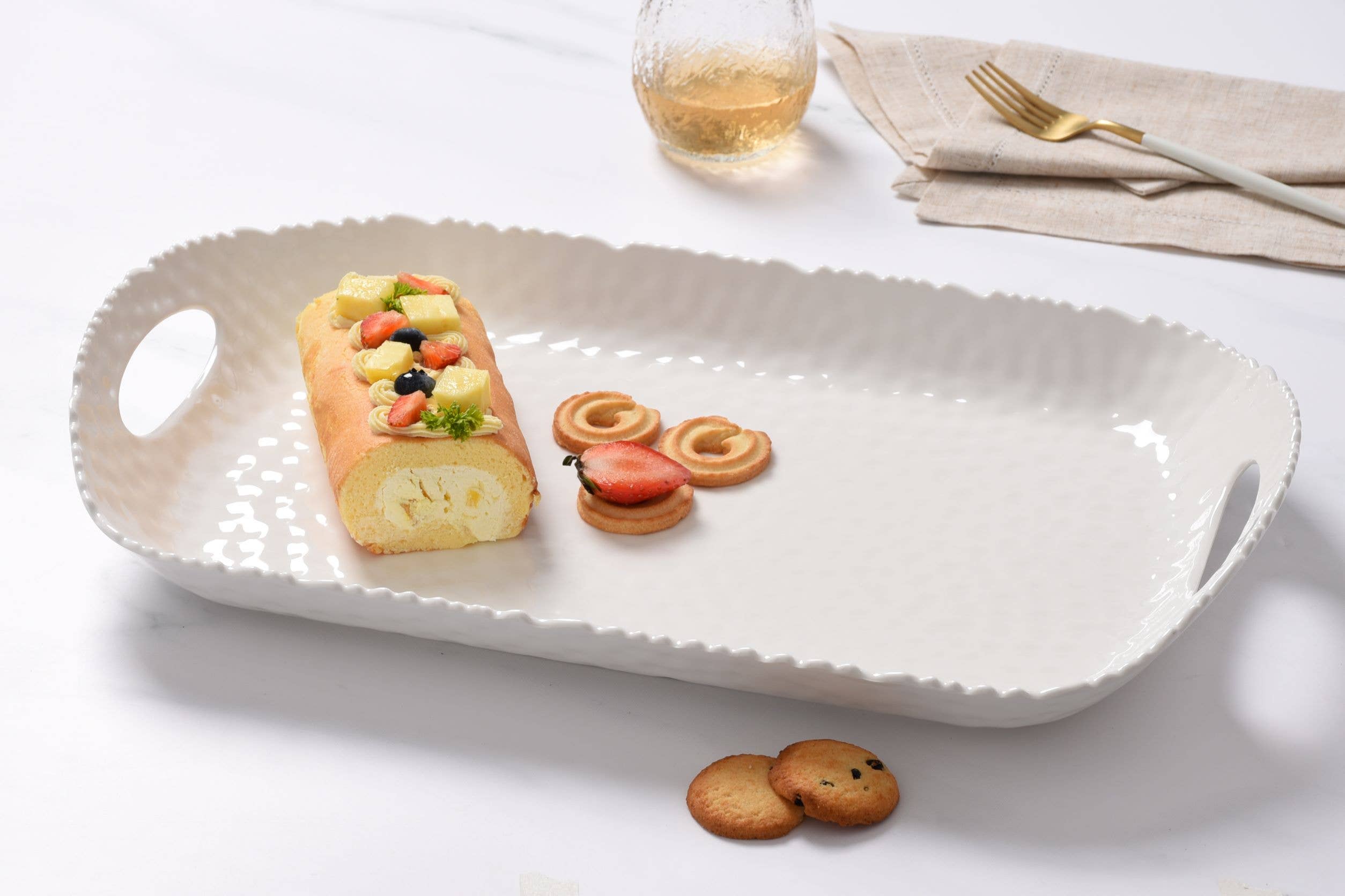 White ceramic platter with a dessert roll, cookies, and a glass of white wine on a light surface.