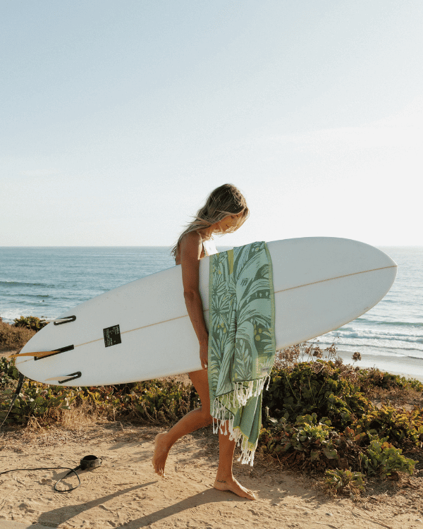 Person walking on a beach with a surfboard and a green towel draped over their shoulder.