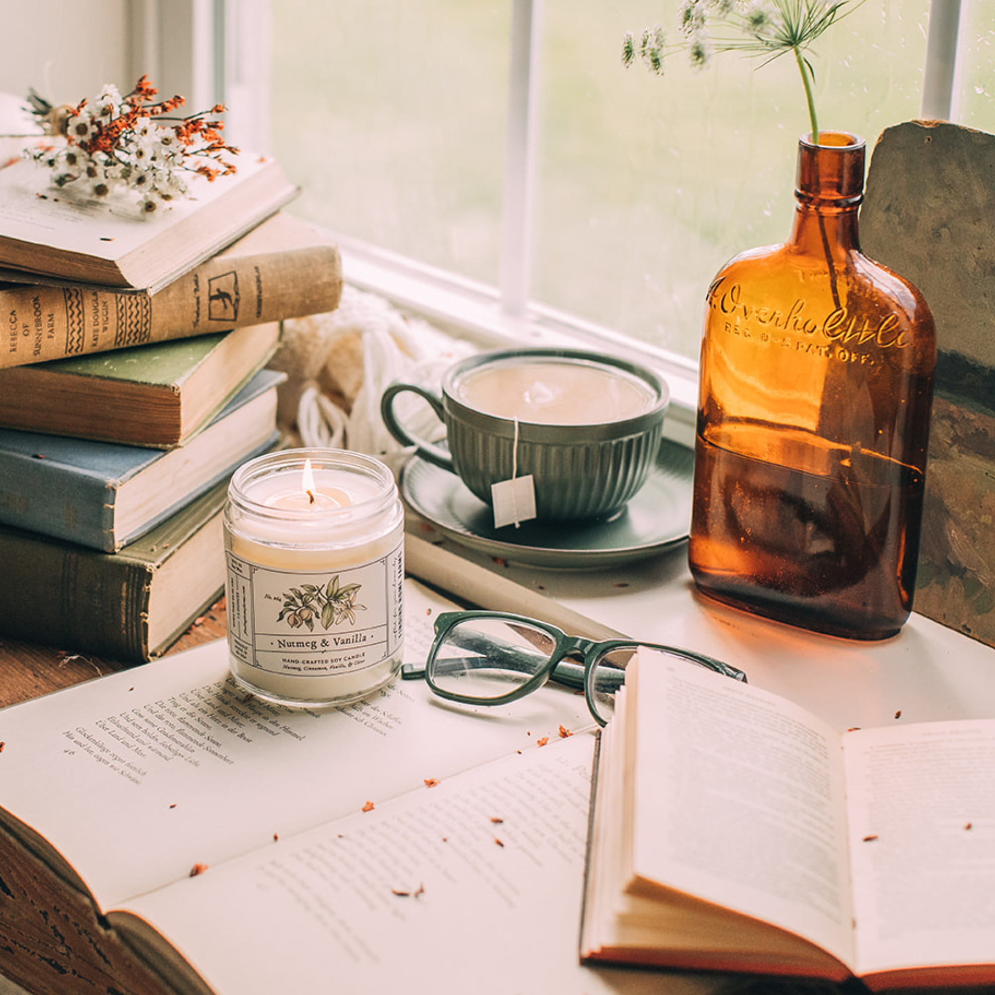 Candle, books, glasses, and a bottle on a table with a window in the background