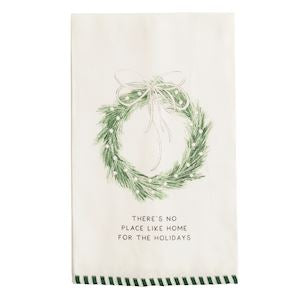 An off-white kitchen tea towel featuring a green pine wreath illustration with a bow and the printed text 'THERE'S NO PLACE LIKE HOME FOR THE HOLIDAYS' above a green and white striped stitched border.