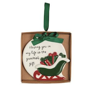 A round white Christmas ornament with a green hanging ribbon, displayed inside a brown gift box. The ornament features a red and green sleigh carrying gifts and the printed message, 'Having you in my life is the greatest gift.