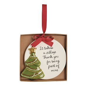 A round ceramic Christmas ornament in a brown gift box with a red ribbon. The ornament is white with black script text reading 'It takes a village thank you for being part of mine' and decorated with a green beaded Christmas tree and a gold star.