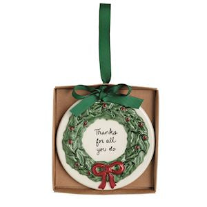 A round ceramic Christmas ornament in a brown gift box, featuring a green wreath design with red berries, a red bow, and the text 'Thanks for all you do' in black script. A green ribbon is tied around the box as a hanger.