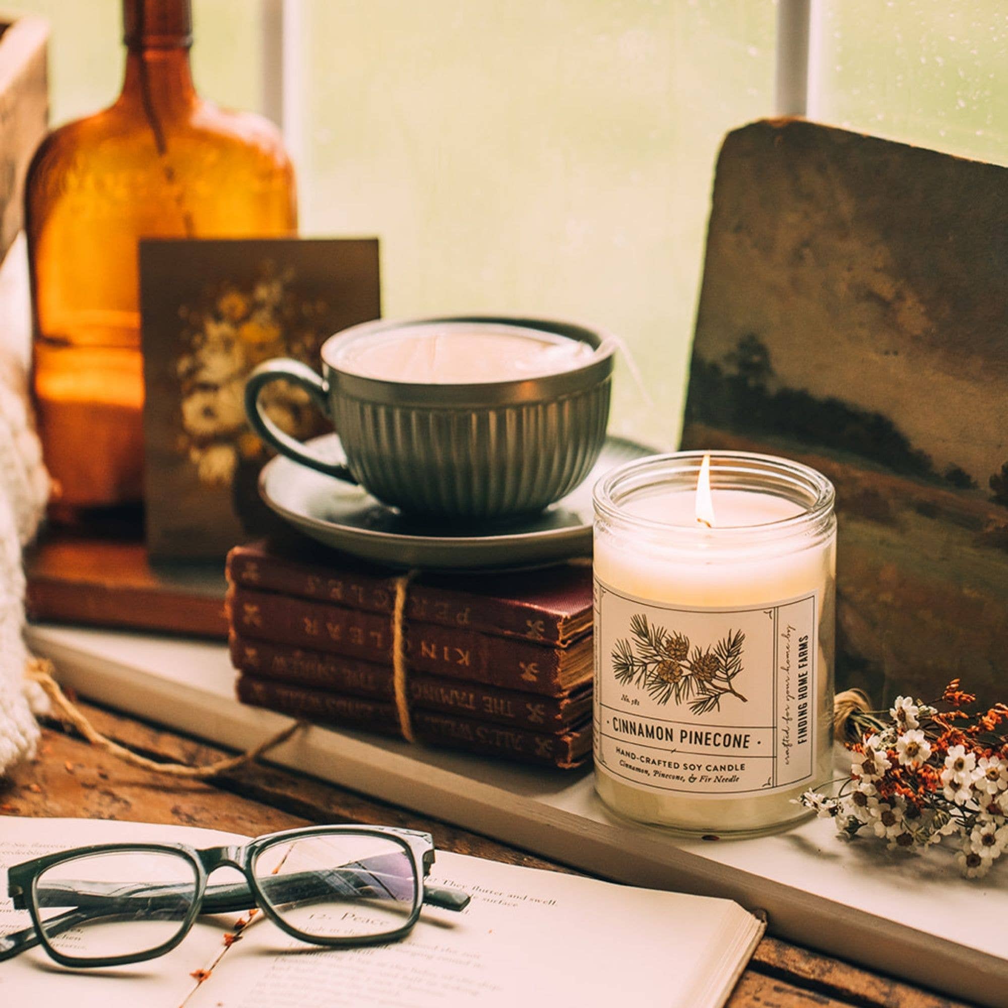 A lit jar candle labeled Cinnamon Pinecone sits on a windowsill beside a cup and saucer, stacked books, eyeglasses on an open book, and dried flowers, with a warm rustic interior background.