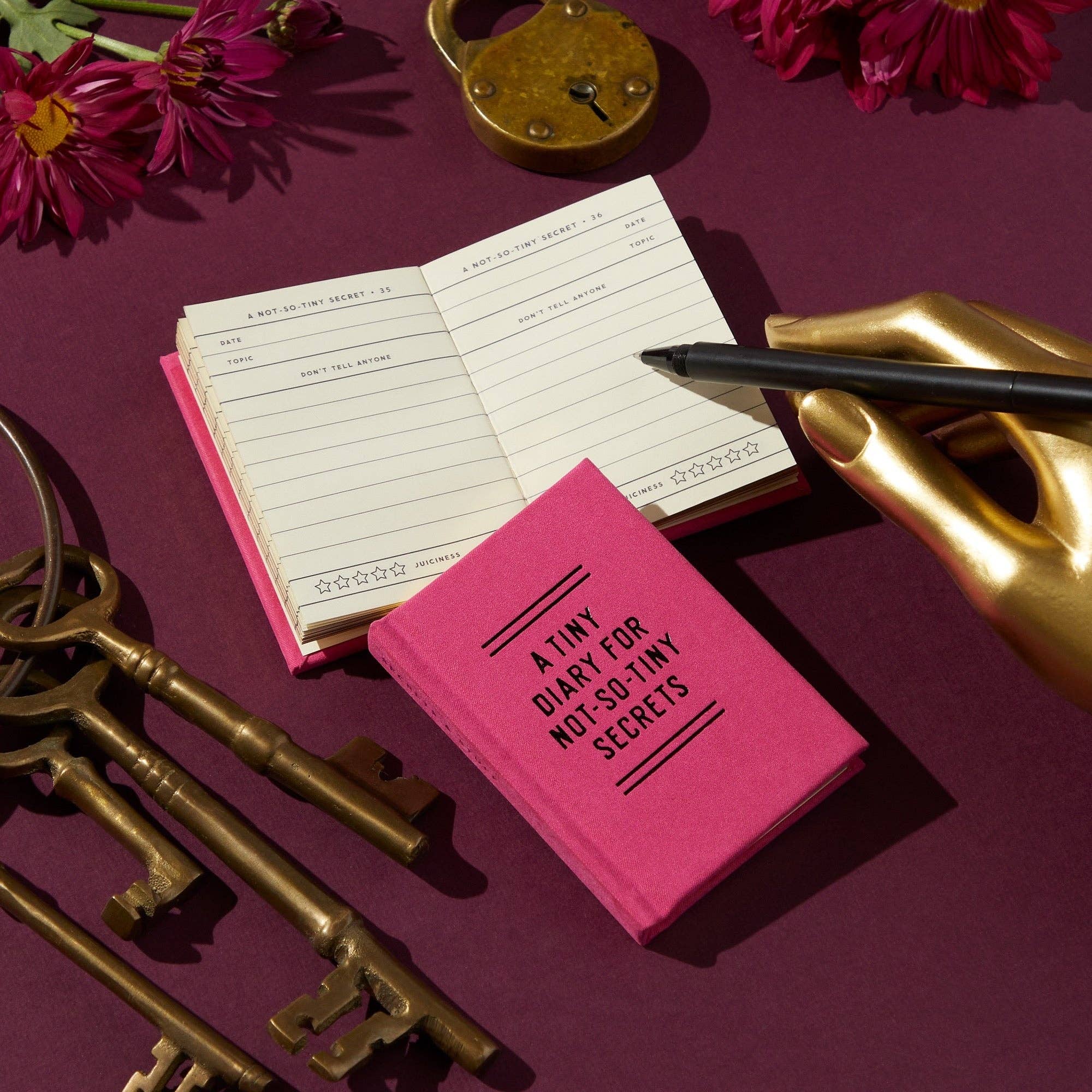 Pink diary with a gold hand holding a pen, surrounded by keys and flowers on a purple background
