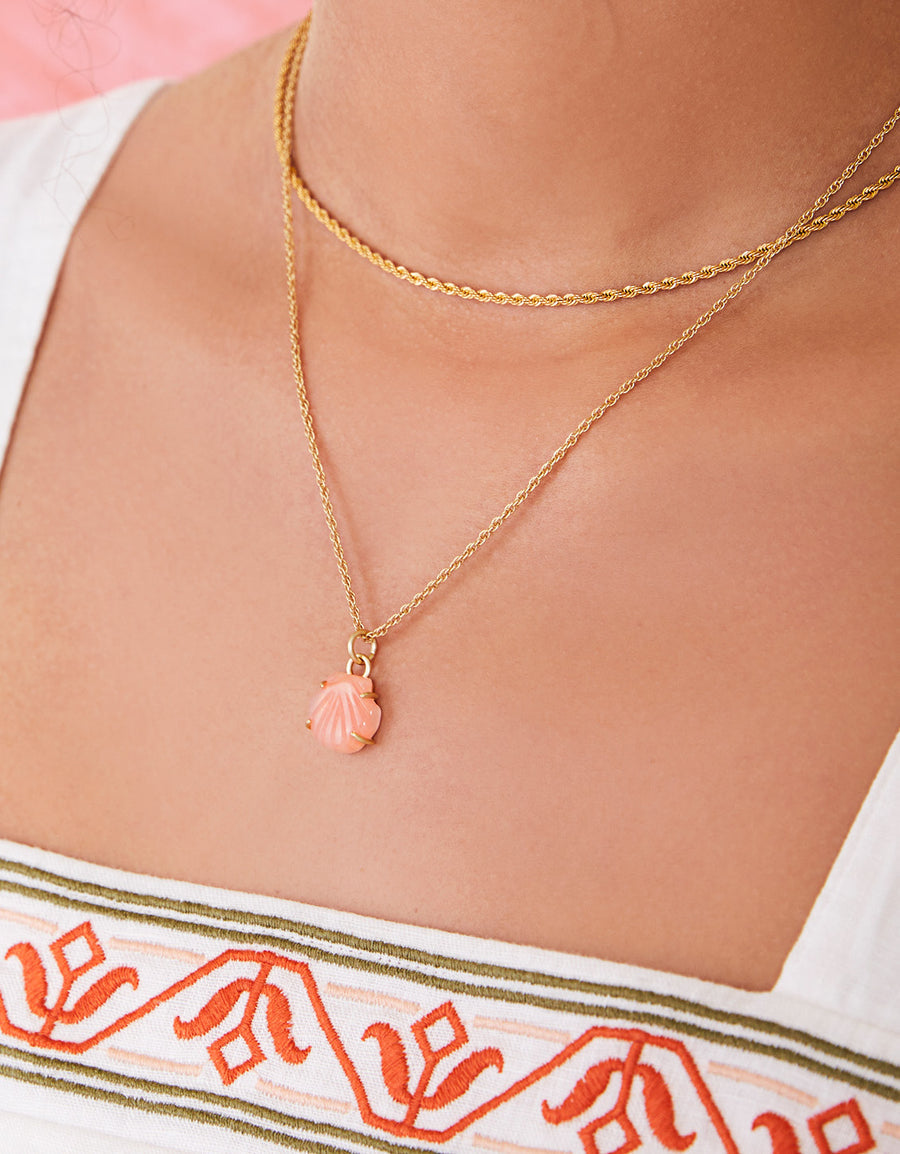 Close-up of a person wearing a gold necklace with a shell pendant, against a blurred background.