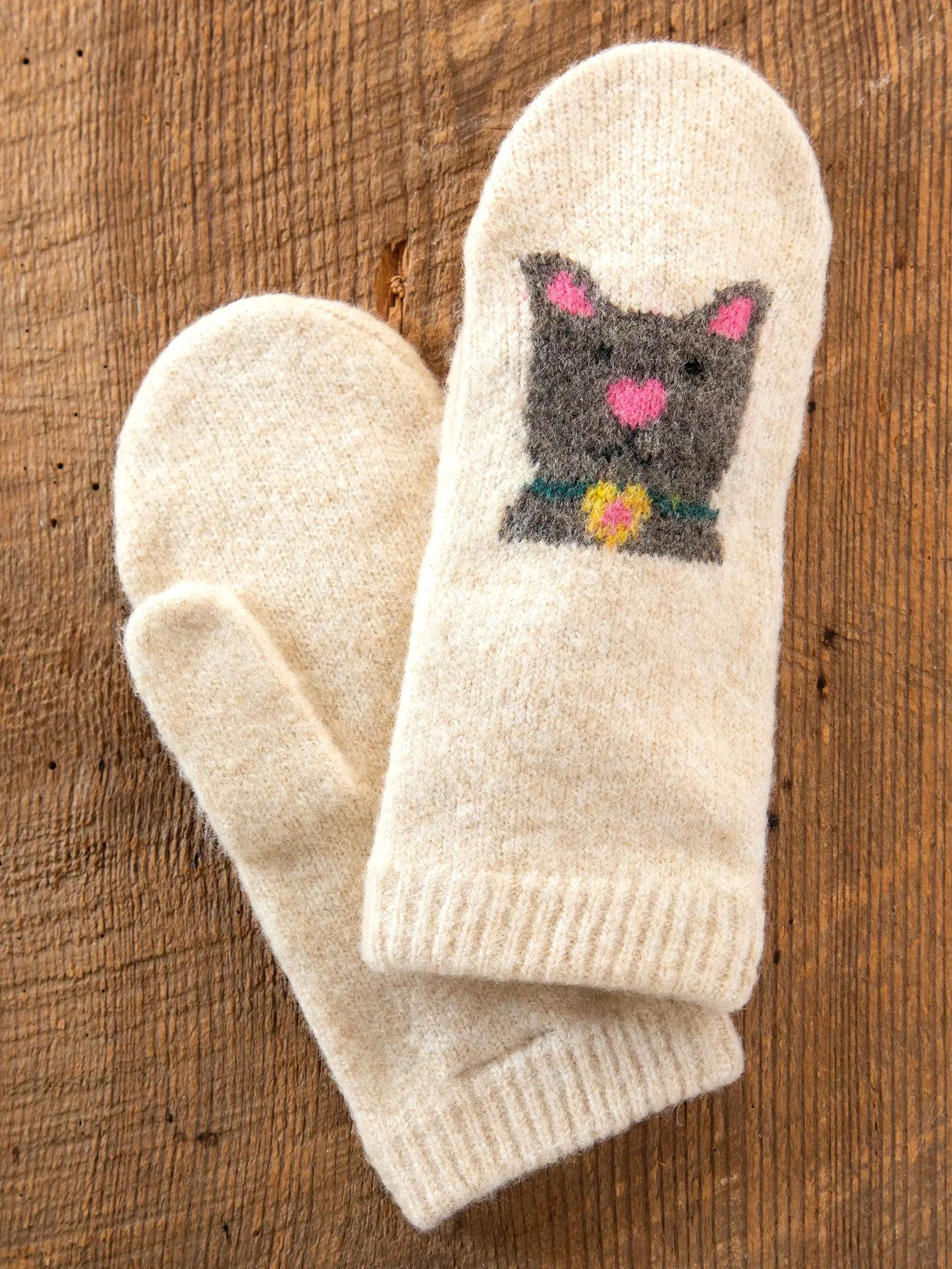 Pair of cream-colored mittens with a cat design on a wooden surface