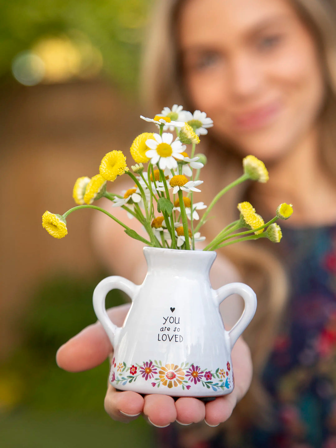 Person holding a small white vase with flowers against a blurred background