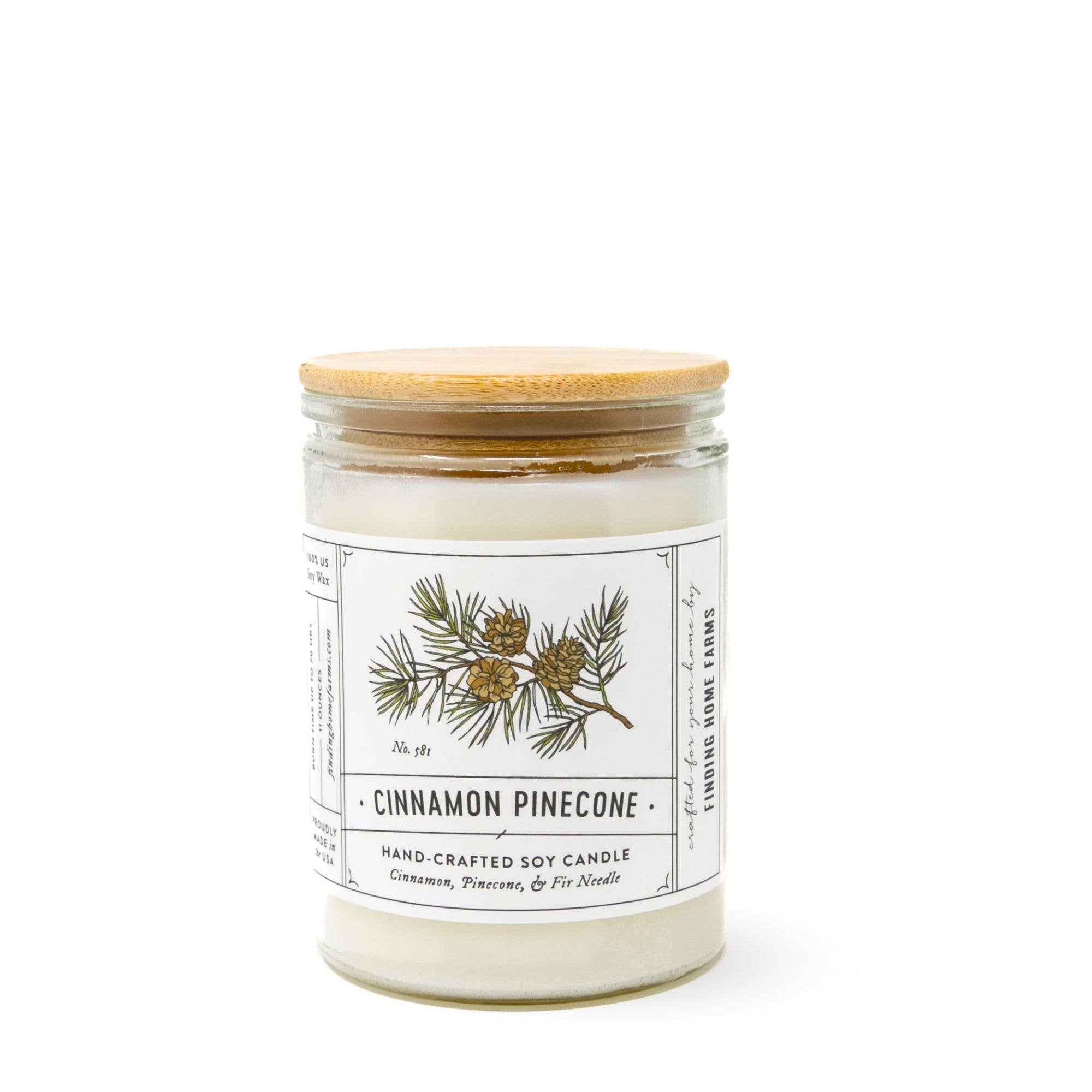 A hand-crafted soy candle in a clear glass jar with a light wood lid, featuring a white label with a pine branch illustration and the scent name 'CINNAMON PINECONE'.
