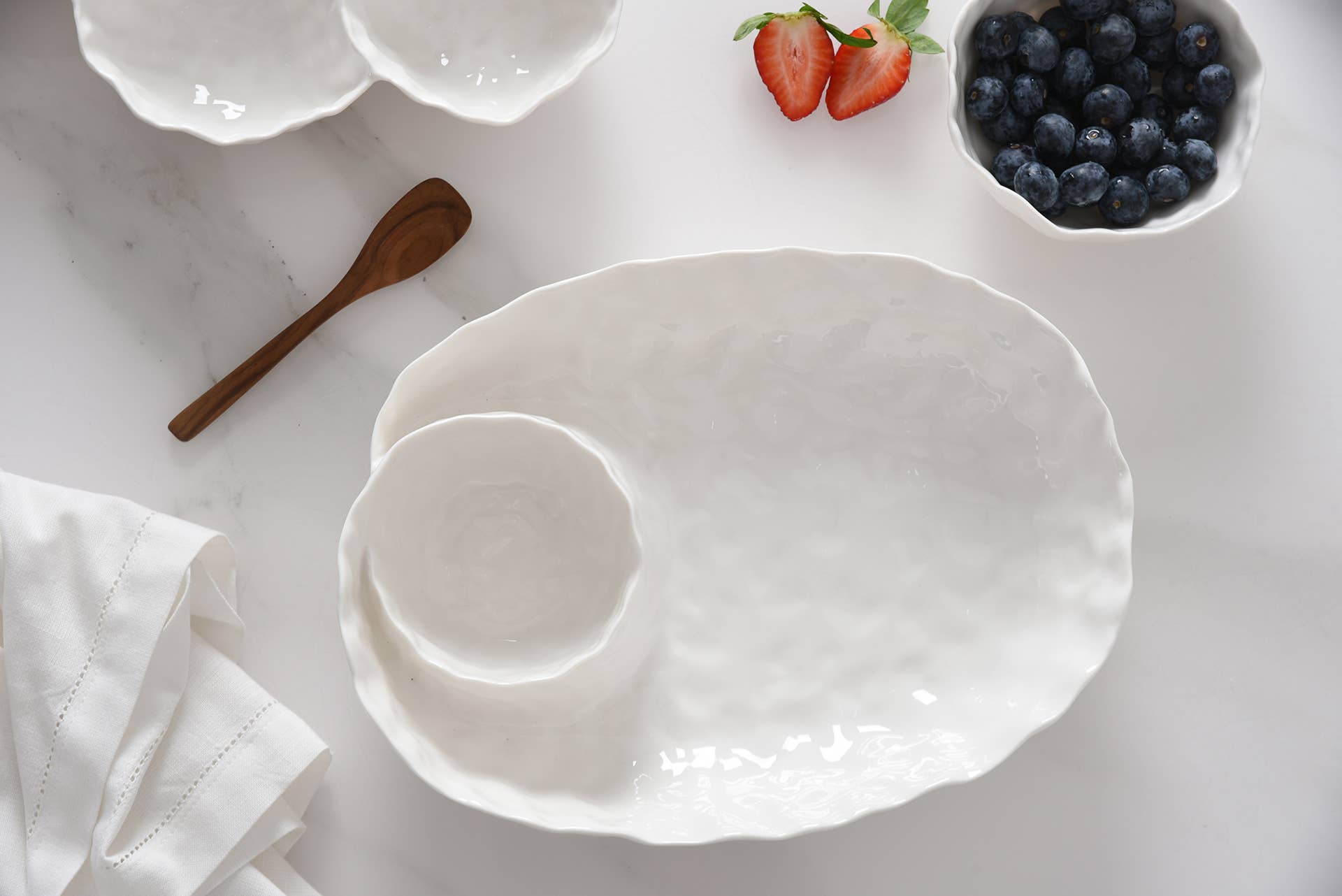 White ceramic platter on a light surface with a wooden spoon and small containers of strawberries and blueberries.