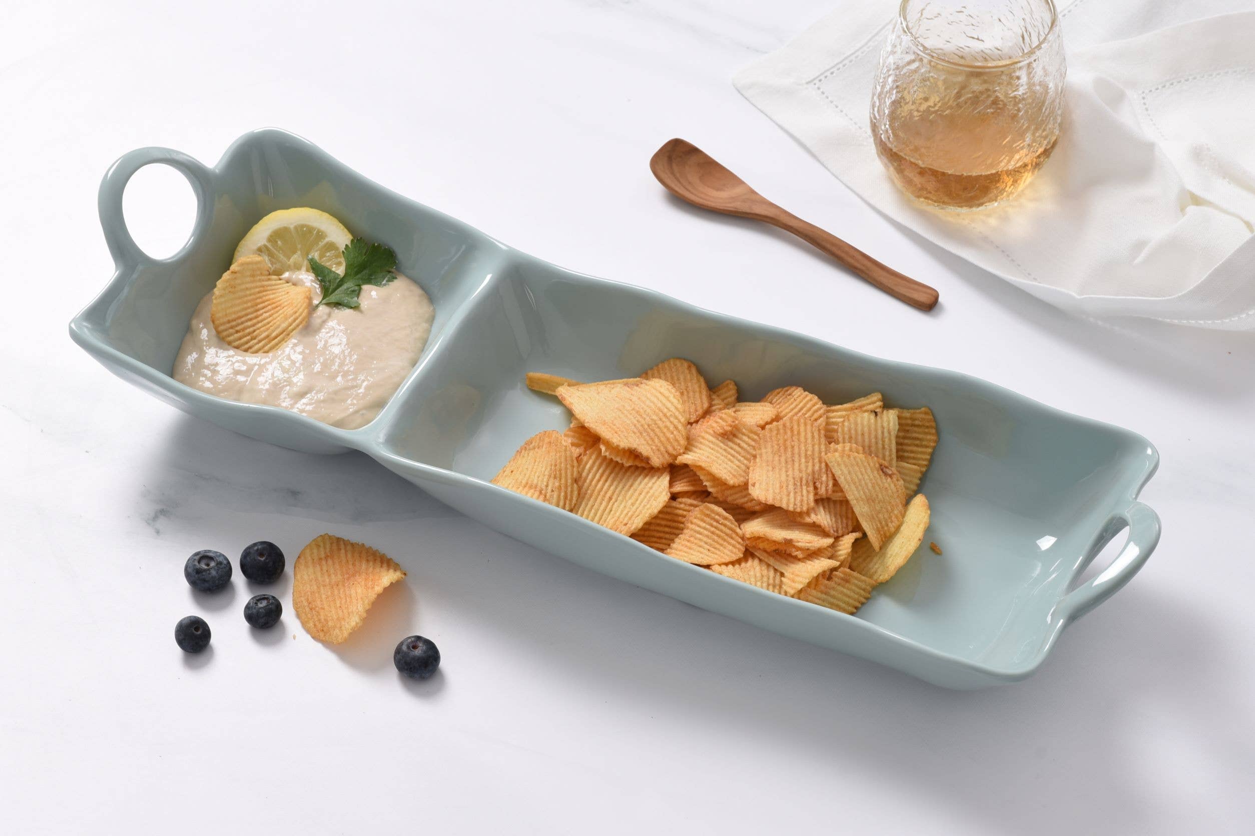 Rectangular ceramic dish with compartments containing chips, dip, and fruit on a white surface.