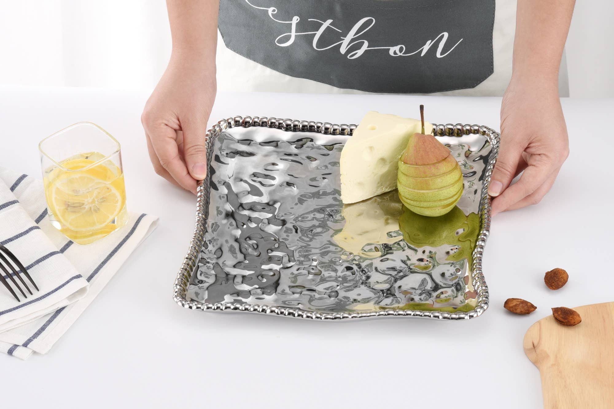 Person holding a decorative tray with a pear and glass of lemonade on a white surface.