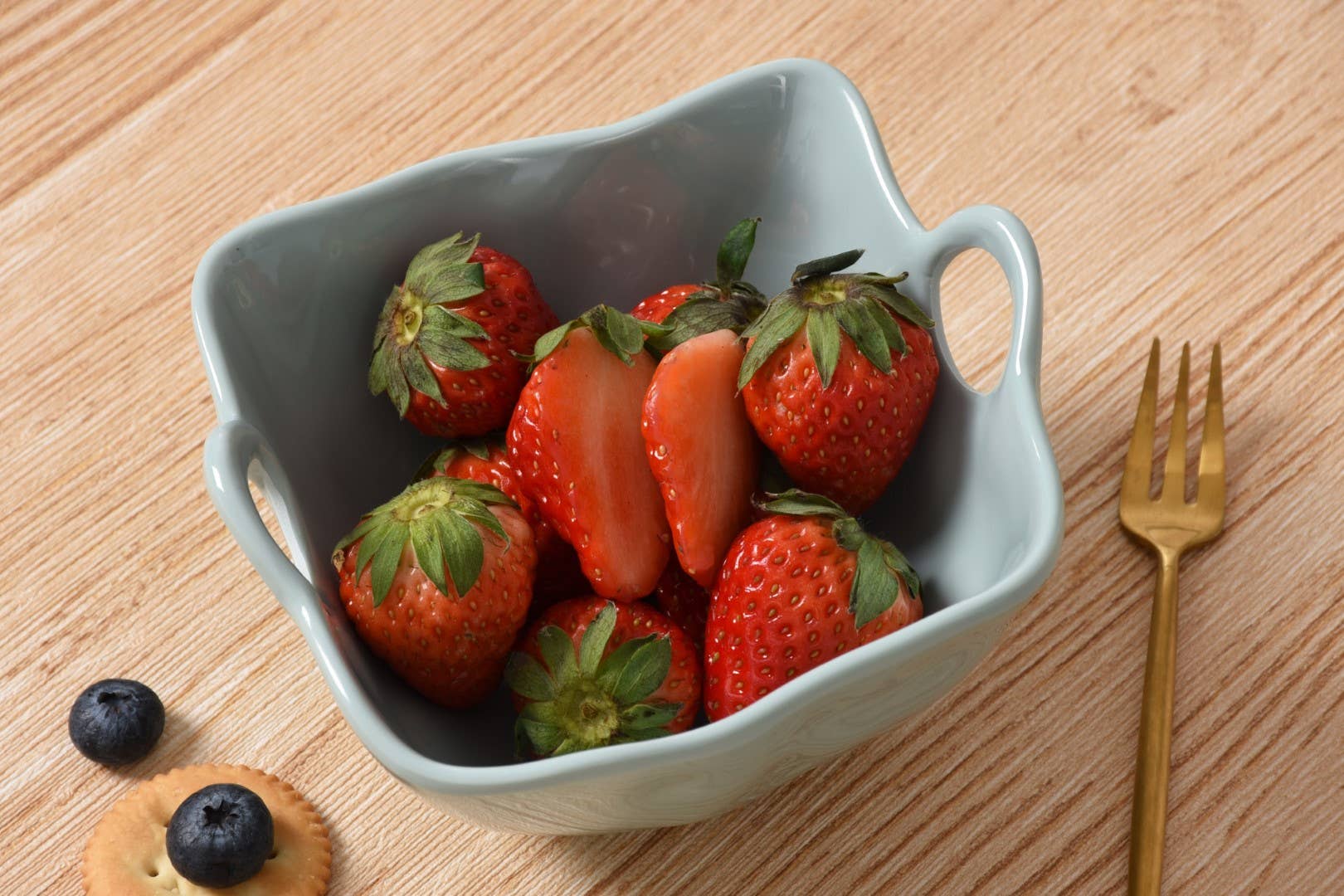 Gray ceramic bowl filled with strawberries on a wooden surface with a gold fork.