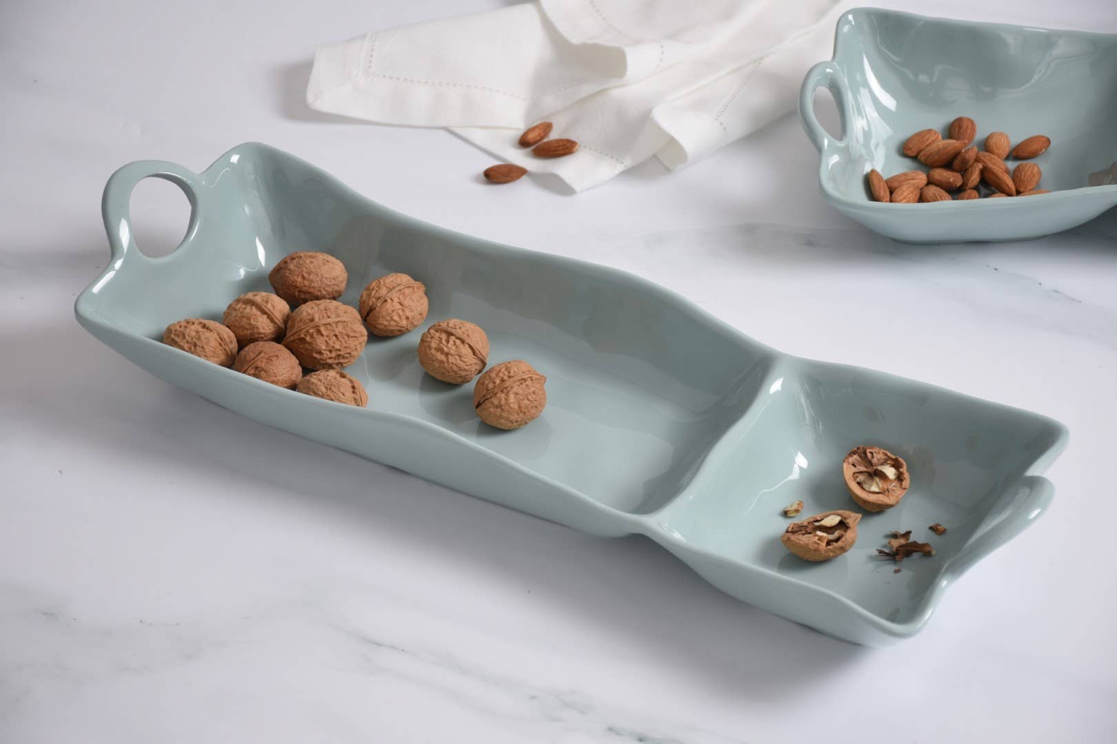 Light blue ceramic dish with handles filled with walnuts on a white marble surface.